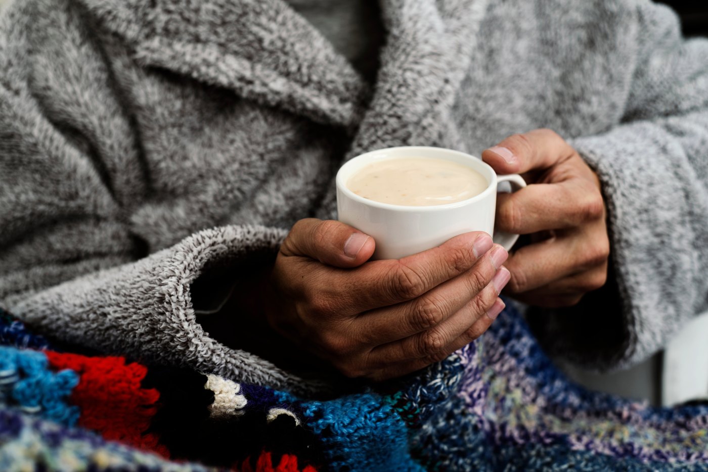 closeup of an ill caucasian man at home, wearing a fluffy gray house robe and wrapped in a colorful knitted blanket, about with warming up with a cup of hot soup